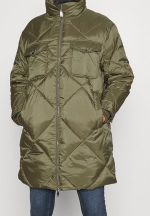 Tikkitekoinen oliivinvihreä pitkä toppatakki, jossa on etuvetoketju, kaksi rintataskua, sivutaskut ja joustavat hihansuut, puettu sinisten farkkujen päälle.