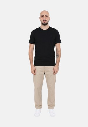 Uomo con testa rasata e barba, indossa una t-shirt nera, pantaloni beige e sneakers bianche, in piedi su sfondo chiaro uniforme, volto verso la visuale frontale.