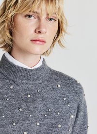 Grauer Pullover mit hohem Kragen, verziert mit verstreuten weißen Perlen und Strass-Applikationen, die eine weiche Strickstruktur zeigen.