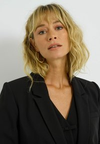 Blazer noir avec col cranté, un seul bouton et légère texture. Le modèle a les cheveux ondulés, mi-longs, et porte des boucles d'oreilles rondes en or.