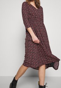 Schwarz-blumiges Kleid mit Dreiviertelärmeln, einem V-Ausschnitt und einer gerafften Taille, das kleine rosa und rote Blumen auf einem strukturierten Stoff zeigt.