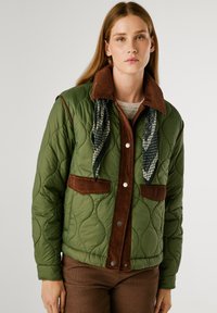 Giacca trapuntata verde con accenti in corduroy marrone, bottoni a pressione e una sciarpa nera a motivi al colletto. Presenta cuciture visibili e tasche.