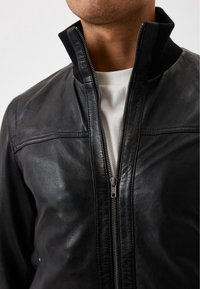 Mężczyzna w czarnej skórzanej kurtce z częściowo rozpiętym zamkiem, pod nią widoczna biała koszula, kołnierz uniesiony.