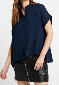 Camicia azzurra navy a maniche corte con bottoni, caratterizzata da un orlo curvo e una tasca frontale, abbinata a una gonna in pelle nera con zip.