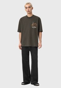 Ciemnozielony oversize T-shirt z wzorzystym nadrukiem i napisem na lewej piersi, zestawiony z luźnymi czarnymi dżinsami i klapkami na nogę.