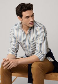 Camisa de cuadros en azul claro, crema y azul oscuro; cuenta con un bolsillo en el pecho. Combinada con pantalones beige, sentado en un banco de madera.