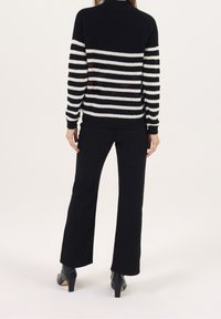 Personne portant un pull rayé noir et blanc, un pantalon noir et des chaussures à talons noires, debout de dos sur un fond uni.