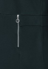 Tissu vert foncé avec une texture lisse, doté d'une fermeture éclair argentée avec un tirette circulaire et d'un détail de couture au-dessus d'une poche zippée.