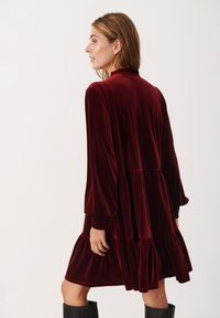 Abito in velluto borgogna con scollatura arricciata, maniche lunghe e fondo a strati. Presenta una vestibilità comoda e una texture morbida.