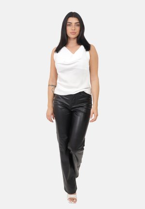 Femme aux cheveux noirs portant un haut sans manches blanc, un pantalon en cuir noir et des talons ouverts blancs, se tenant devant un fond clair.