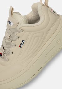 Sneaker beige con tomaia in tessuto testurizzato, punta rotonda e colletto imbottito. Presenta il logo Fila in blu, rosso e bianco sul lato.