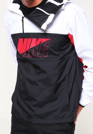 Personne portant une veste Nike noire et blanche avec un logo rouge, debout avec une main dans la poche, visible du cou à la taille.