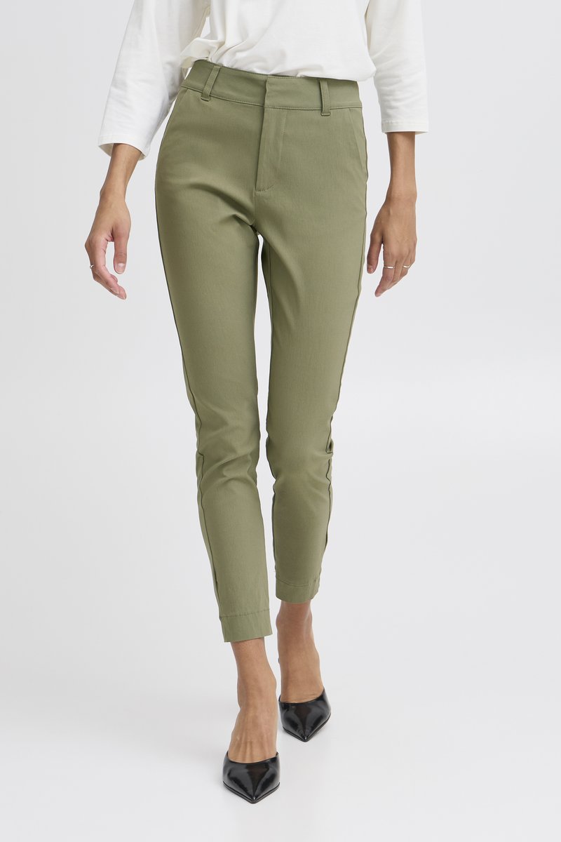 Femme portant un pantalon vert olive ajusté et des talons pointus noirs, avec un haut blanc à manches longues sur un fond uni.