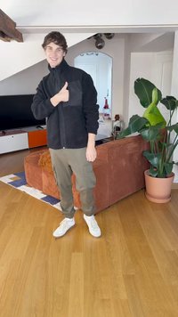Veste en polaire noire zippée avec une poche poitrine, pantalon cargo olive et baskets blanches, dans un environnement intérieur avec une plante et un canapé.