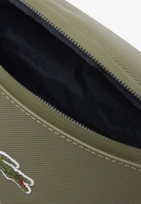 Borsetta in stoffa verde testurizzata con chiusura a zip, con un interno nero e un logo di coccodrillo ricamato bianco sulla parte frontale.