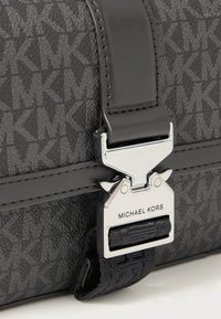Zwarte tas met een patroon van het MK-monogram, voorzien van een zilveren sluiting met de merknaam, leren accenten en een gestructureerde stoffen afwerking.