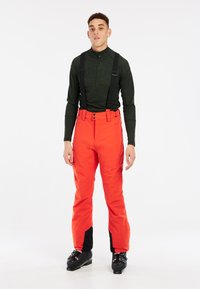 Pantalons de ski rouges avec bretelles noires ajustables, coupe ajustée, renforcement dans les sections inférieures, portés avec un haut à manches longues vert foncé et des bottes de ski noires.