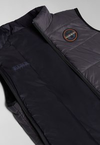 Gilet rembourré noir avec un col haut, doté d'une fermeture éclair avant, d'une texture matelassée et d'un patch logo rond en orange et noir sur la poitrine.