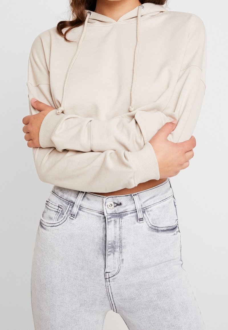 Personne portant un sweat à capuche court beige avec cordons et un jean gris clair taille haute, bras croisés sur la poitrine.