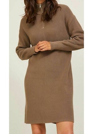 Kobieta ubrana w długą, brązową sukienkę dzianinową z prążkowanej dzianiny z półzamkiem przy kołnierzu, z dłońmi splecionymi na wysokości talii.