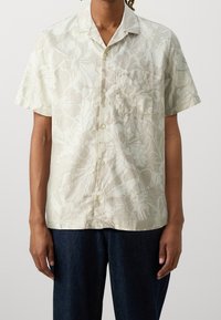 Korte mouwen shirt in lichtbeige stof met subtiele bloemenpatronen. Heeft een kraag en een borstzak, gecombineerd met donkere denimbroeken.