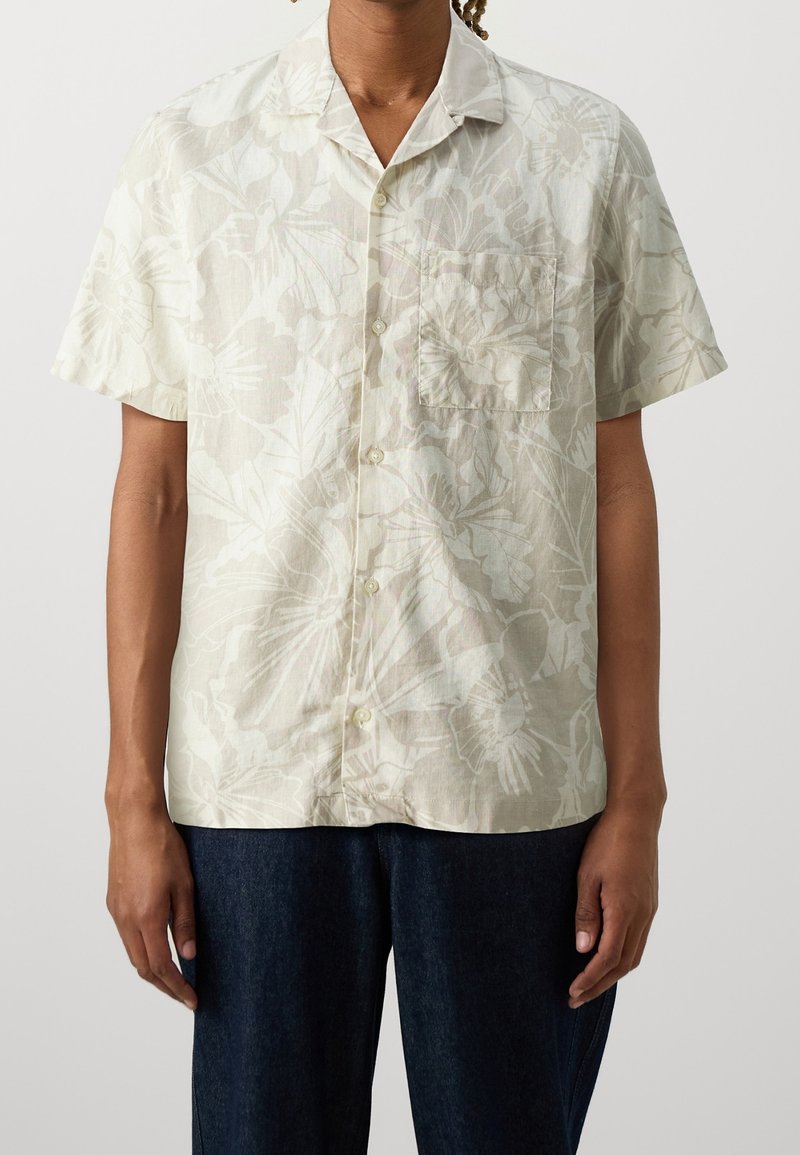 Korte mouwen shirt in lichtbeige stof met subtiele bloemenpatronen. Heeft een kraag en een borstzak, gecombineerd met donkere denimbroeken.