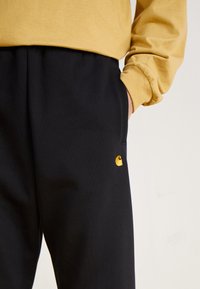Black sweatpants with elastic waistband, side pockets, featuring a small yellow logo on the right leg. Cotton material, soft texture.