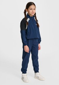 Mörkblå träningsdräkt med svarta detaljer, zip-up jacka, elastiska ärmslut och matchande byxor. Vita hög-top sneakers med grå detaljer.