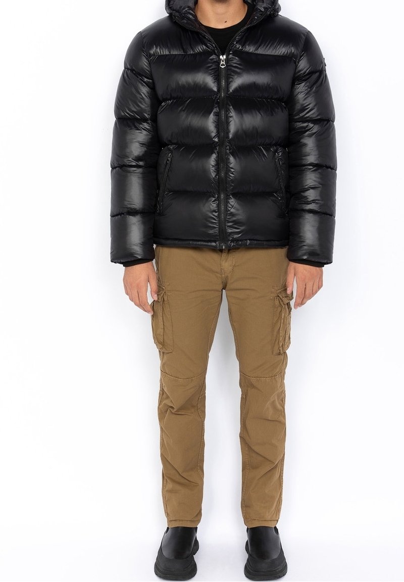 Personne portant une doudoune noire avec fermeture éclair à l’avant, un pantalon cargo beige avec poches latérales et des bottes noires, debout devant un fond blanc.