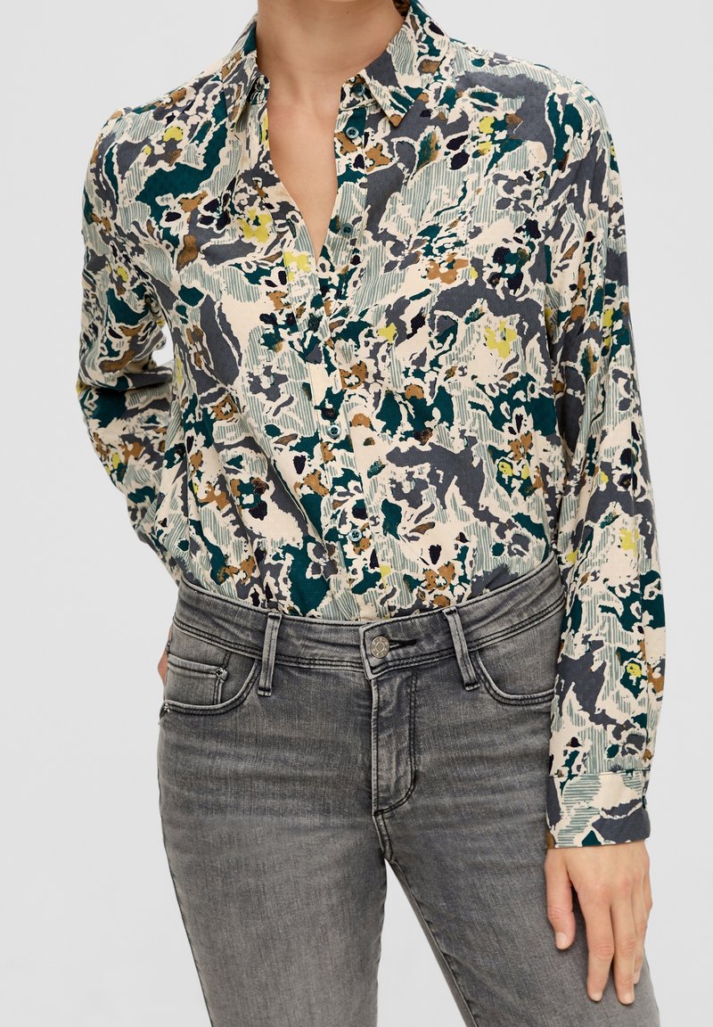 Camicia button-up con stampa floreale multicolore su sfondo crema, caratterizzata da un colletto aperto e maniche lunghe, abbinata a jeans in denim grigi.