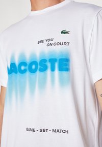 Vit bomull t-shirt med en blå gradient, "LACOSTE" i feta bokstäver, och "SEE YOU ON COURT" ovanför, med en grön logotyp på bröstet.