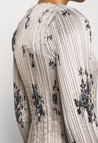 Dettaglio di un top in tessuto beige plissettato con stampa floreale nera sulla parte superiore della schiena e sulla spalla di una persona.