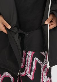 Zwarte jas met een gladde buitenkant, ritsluiting en drukknoopjes op de binnenvoering. De binnenlaag is contrast zwart fleece.