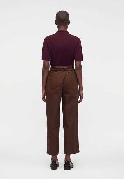 Personne debout de dos portant une chemise bordeaux à manches courtes, un pantalon marron taille haute avec une ceinture et des chaussures foncées sur fond blanc.
