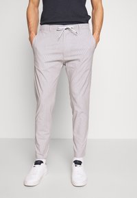 Muž v lehkých šedých slim-fit kalhotách s šňůrkovým pasem, černém tričku a bílých teniskách na jednobarevném pozadí.