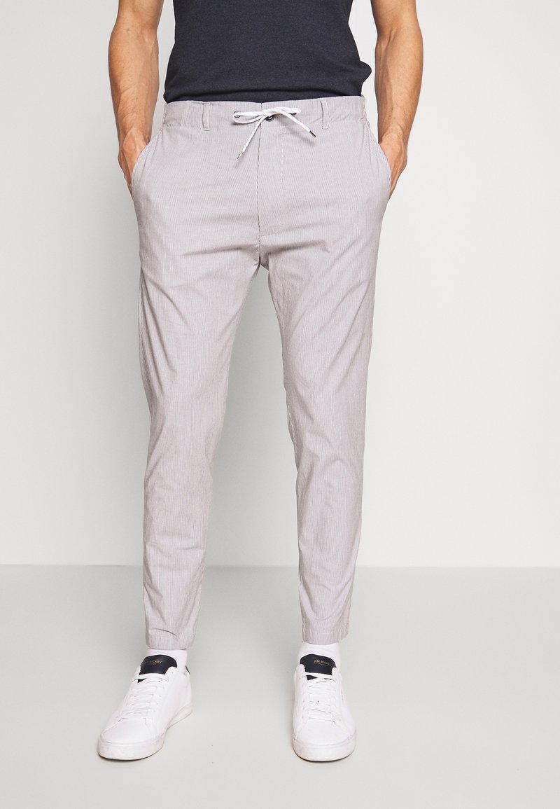 Muž v lehkých šedých slim-fit kalhotách s šňůrkovým pasem, černém tričku a bílých teniskách na jednobarevném pozadí.