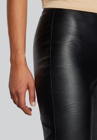 Leggings de couro preto com textura suave, mostrando costuras visíveis e um ajuste justo ao longo da perna, combinados com um top beige liso.