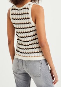 Stickad tanktop i krämfärg med zigzagmönster i svart och brunt, med rund halsringning och ärmlös design. Buren med ljusgrå jeans.