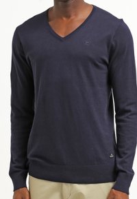 Homme portant un pull long à col V bleu marine et un pantalon beige clair, se tenant contre un fond blanc.