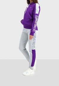 Felpa viola con accenti e branding bianchi, abbinata a pantaloni sportivi grigi con pannelli viola e dettagli del logo. Sneakers bianche.