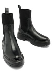 Paire de bottines Chelsea en cuir noir avec semelles épaisses et panneaux en maille côtelée autour de la cheville, sur fond blanc.