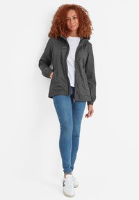 Woman with curly red hair wearing a gray star-patterned zip-up jacket, white shirt, blue skinny jeans, and white sneakers, standing against white wall.