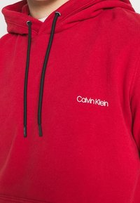 Sudadera roja de algodón con un cuello acanalado. Cuenta con cordones negros y un logo blanco de "Calvin Klein" en el pecho. Textura suave.