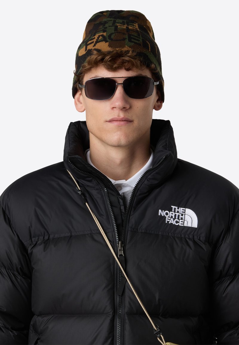 Giubbotto imbottito nero con collo alto, logo bianco, berretto lavorato a maglia marrone e verde con motivo, e occhiali da sole scuri. Presenta una chiusura a zip.
