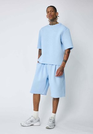 Homme avec des tatouages portant un t-shirt oversize bleu clair et un short, des chaussettes blanches et des baskets argentées, debout les mains dans les poches sur un fond uni.