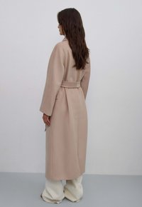 Beige lange jas van zachte stof, met een ceintuur in de taille, brede mouwen en een rechte snit, gecombineerd met neutrale wijde broek.