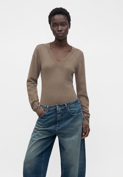 Femme debout, les mains dans les poches, portant un pull marron à col en V et un jean bleu ample sur un fond blanc uni.