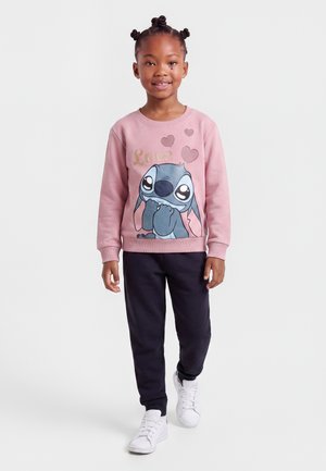 Fille souriante aux cheveux tressés porte un sweat-shirt rose avec un personnage de dessin animé et des cœurs, pantalon noir et baskets blanches, debout sur fond blanc.