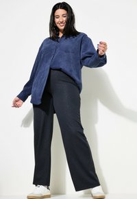 Marineblauwe suedine oversized blouse, twee borstzakken; zwarte broek met verticale strepen en wijde pijpen; witte sneakers met tan accenten. Eenvoudig en gestructureerd ontwerp.