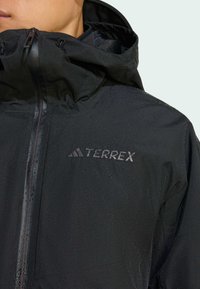 Czarna, wodoodporna kurtka z wysokim kołnierzem, zamkiem z przodu i logo "TERREX". Charakteryzuje się gładką teksturą z widocznymi kroplami wody.